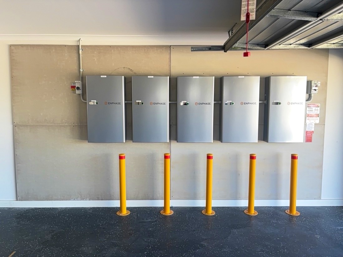 Five Enphase IQ batteries in a row on a garage wall, each protected by a yellow bollard