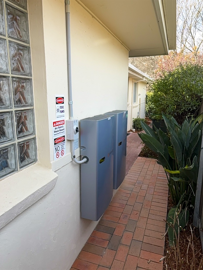 Two Enphase batteries mounted on a cream wall beside a brick path
