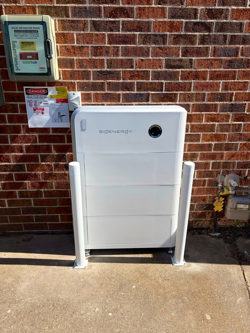 Sigenergy battery with white bollards on a red brick wall beside an Enphase solar board