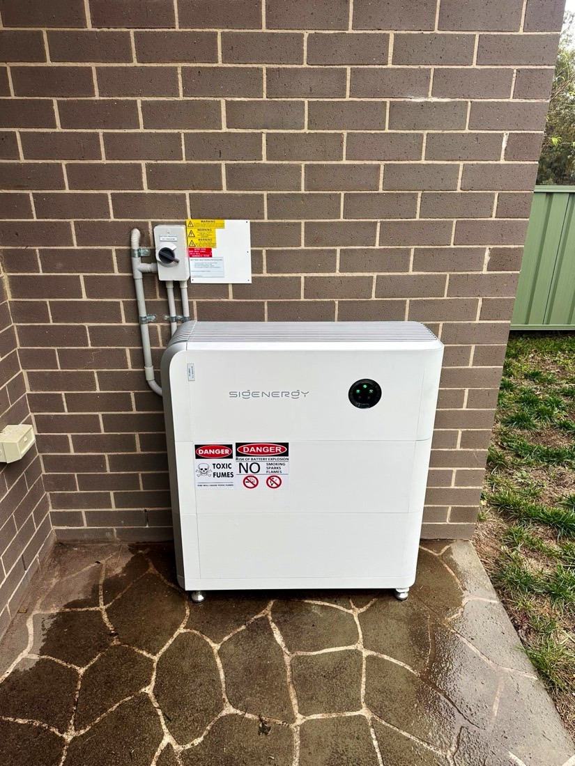 Sigenergy battery on a paved corner beside a brown brick wall