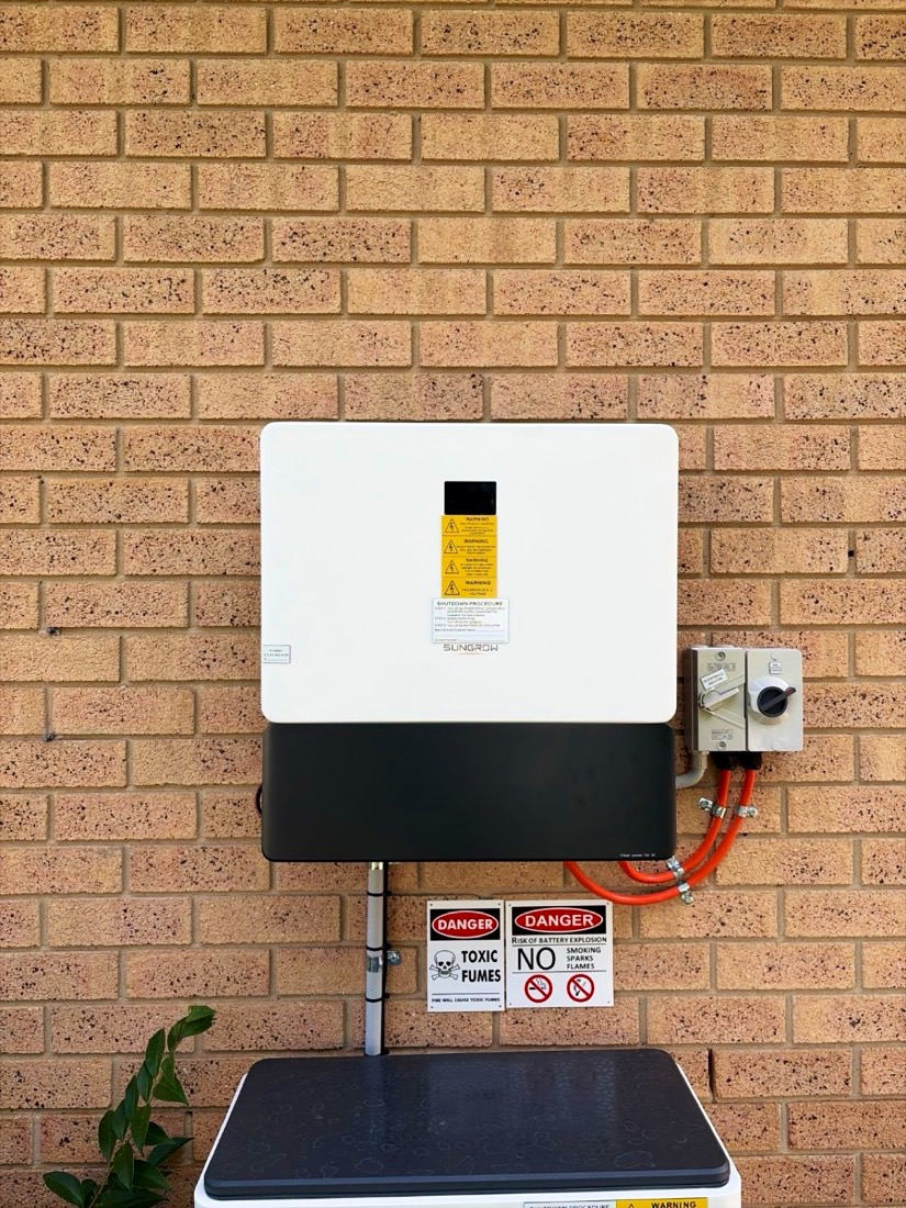 White Sungrow hybrid inverter mounted on a brick wall above a battery