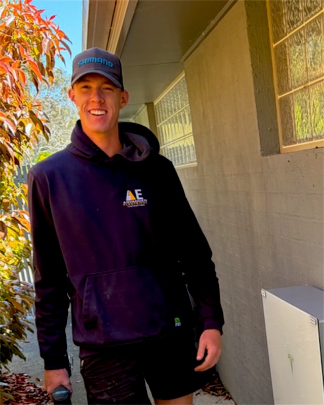 Hayden smiling beside a Tesla Powerwall install, holding a drill