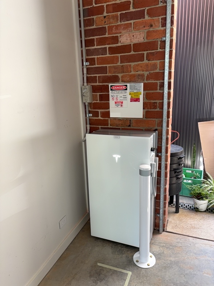 Tesla Powerwall in a carport corner with isolator, signage and a white protective bollard