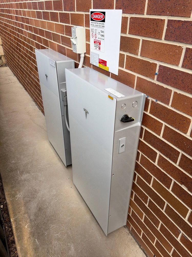 Tesla Powerwall 3 and Expansion unit side by side along a red brick wall with isolator and signage