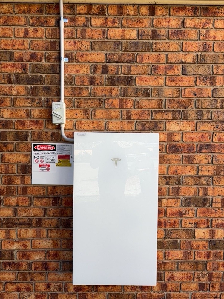 White Tesla Powerwall wall-mounted on a red brick wall with isolator and conduit run