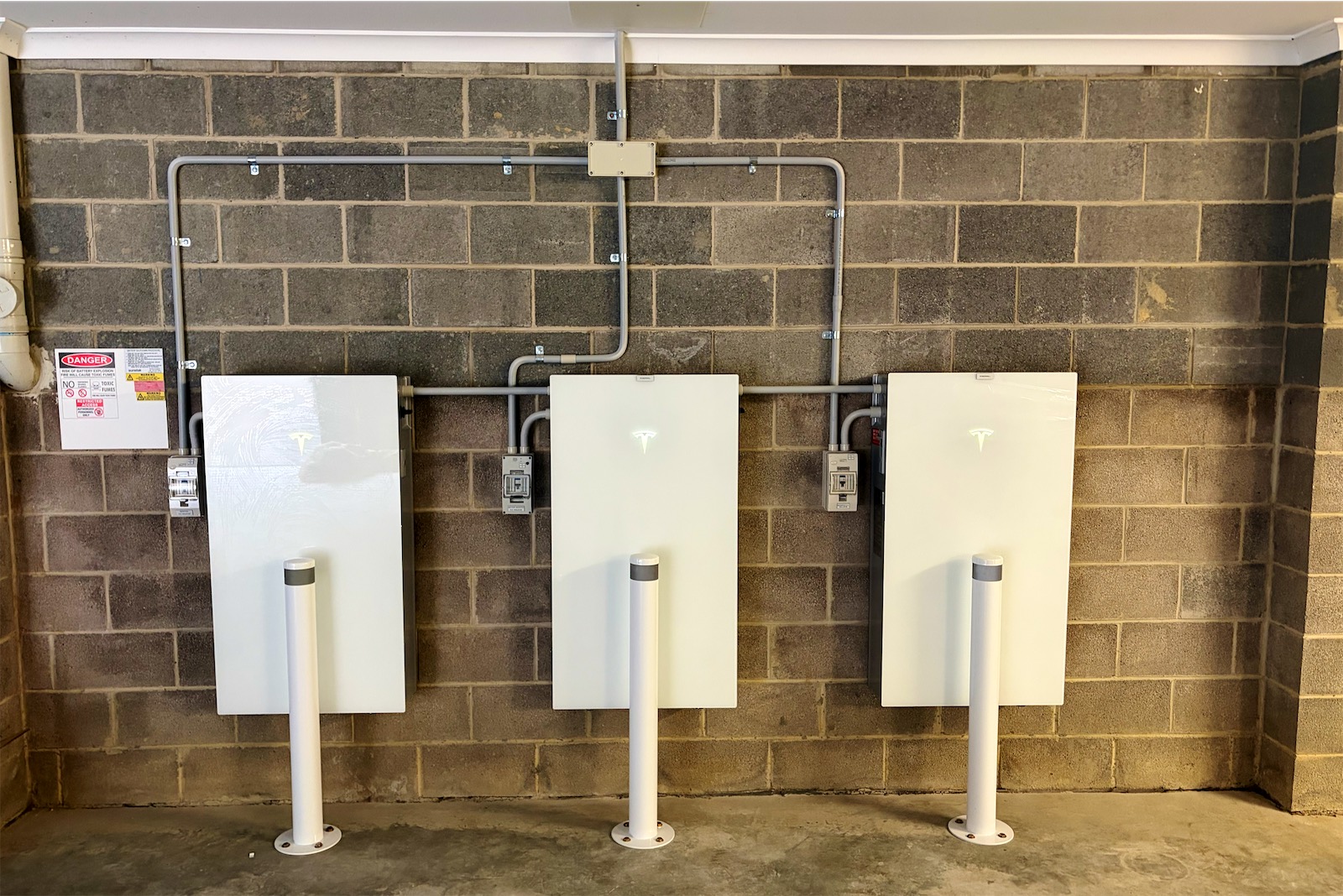 Three Tesla Powerwall units on a block garage wall, each with its own isolator, conduit run and white bollard